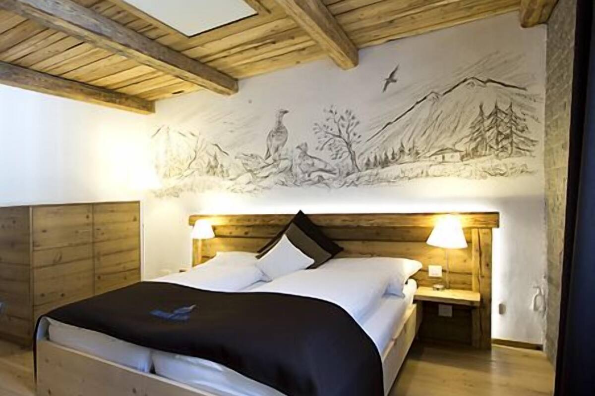 A bedroom with a wooden ceiling, a bed with white bedding, two bedside lamps, and a mural on the wall depicting a landscape scene.