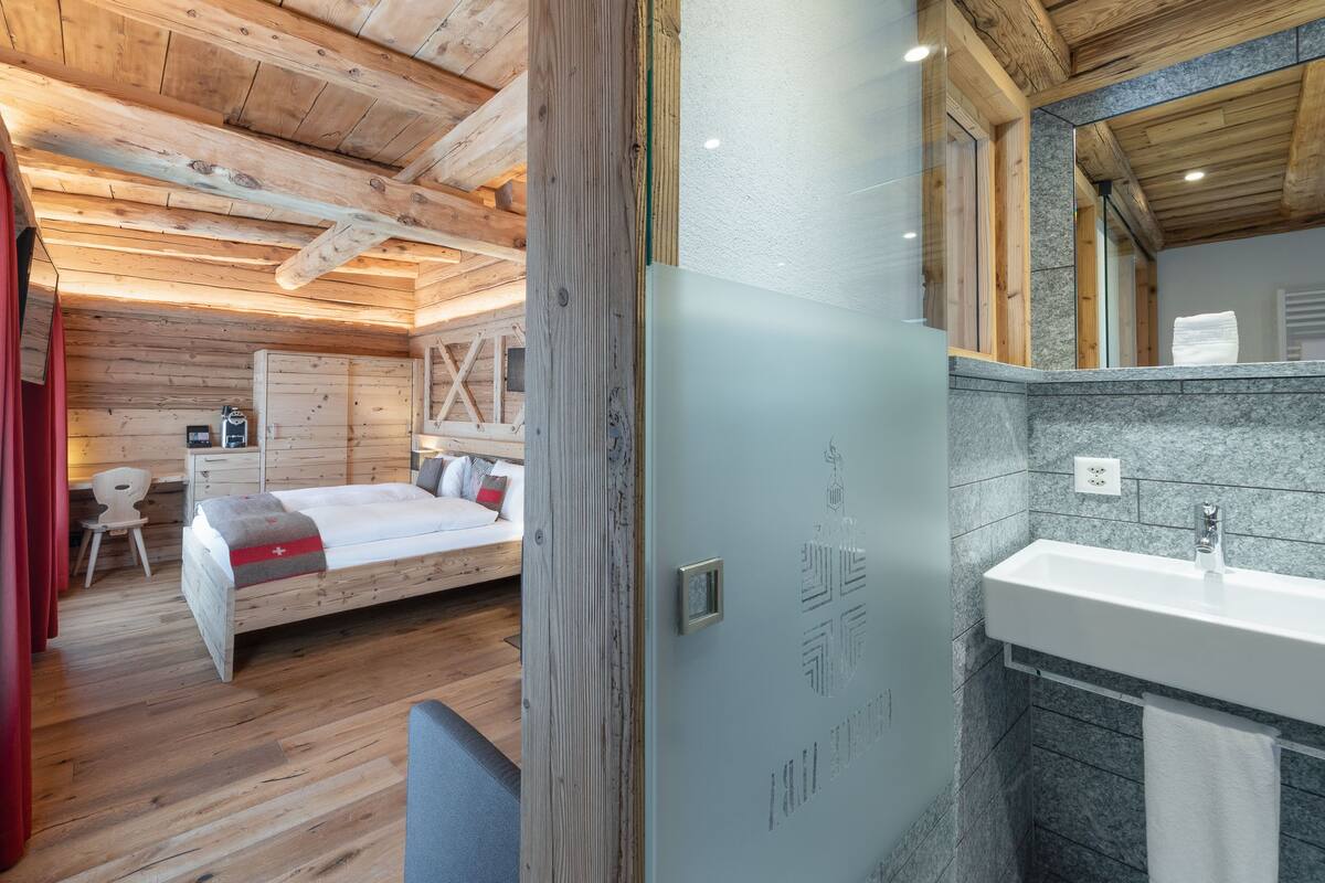 A wooden cabin-style room with a bed, a small desk, and a bathroom with a sink and mirror.