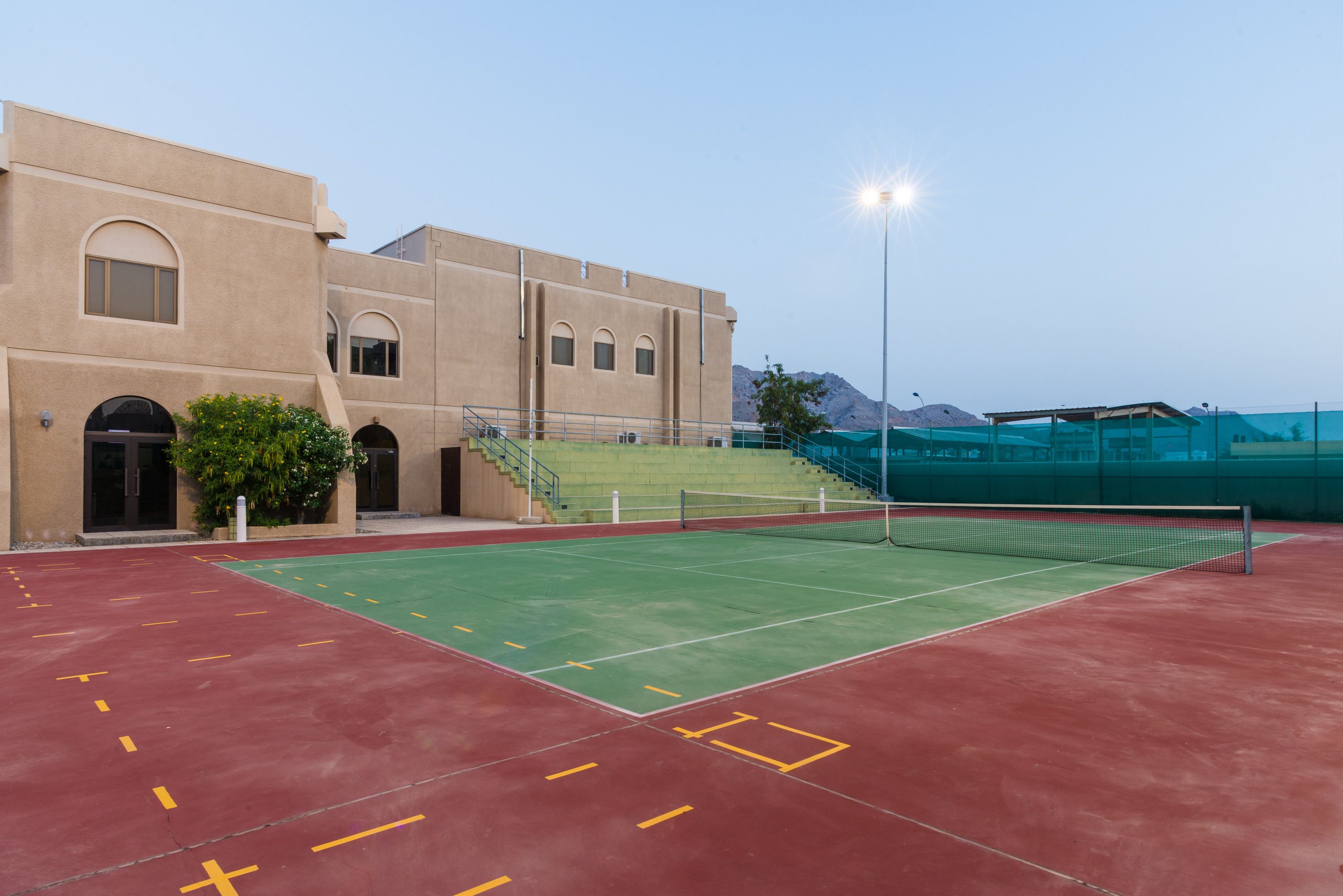 An outdoor tennis court adjacent to a building with arched windows and a staircase leading to a green area.