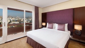 A hotel room with a large bed, a nightstand, a lamp, and a view of a cityscape through a sliding door. - Al Falaj Hotel (Muscat)