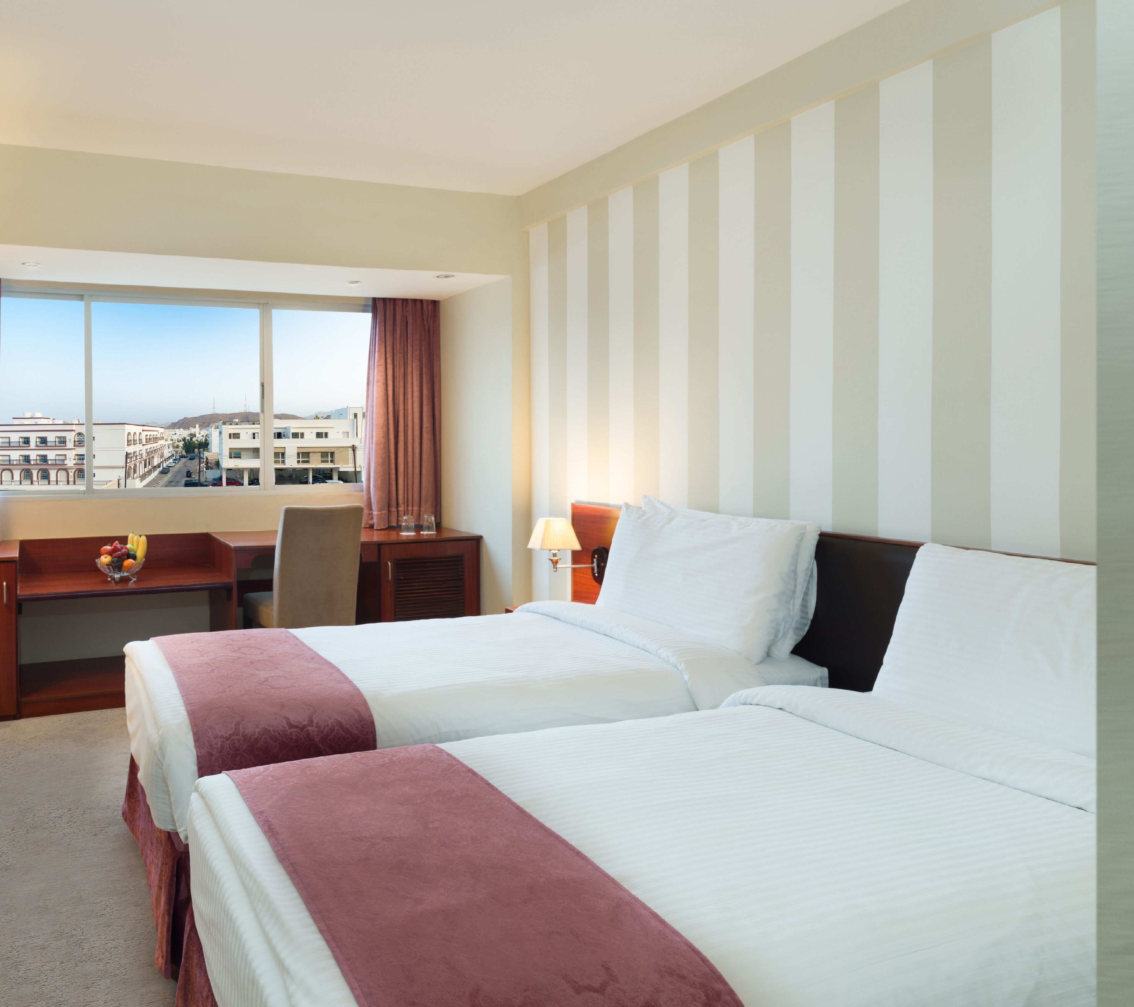 A hotel room with two beds, a desk, a chair, a lamp, and a view of the city through large windows.