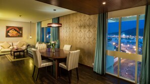 A dining area with a wooden table and chairs, a hanging light fixture, and a view of a cityscape through large windows. - Al Falaj Hotel (Muscat)