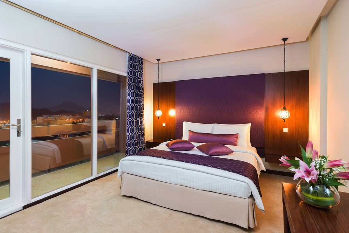 A modern bedroom with a large bed, a view of a cityscape through a sliding glass door, and a decorative wall panel.