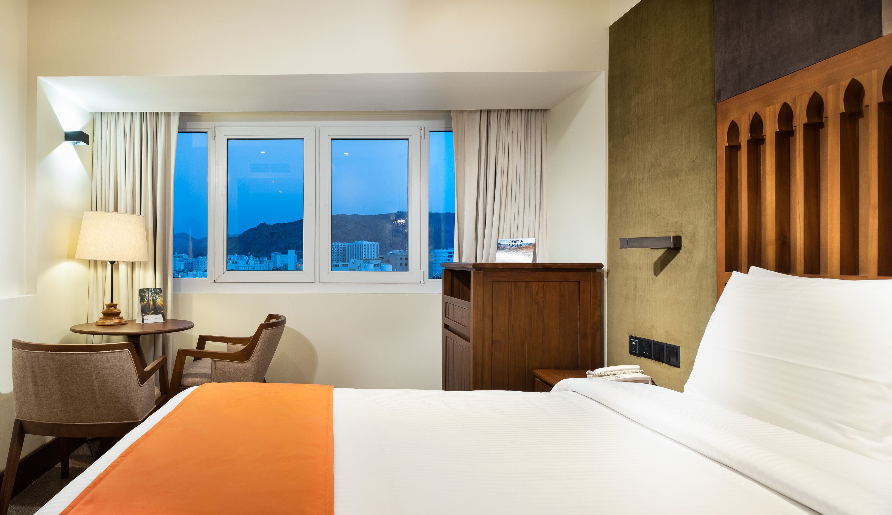 Habitación de hotel con cama, escritorio con lámpara, silla, ventana con cortinas y vista a un paisaje urbano al anochecer.