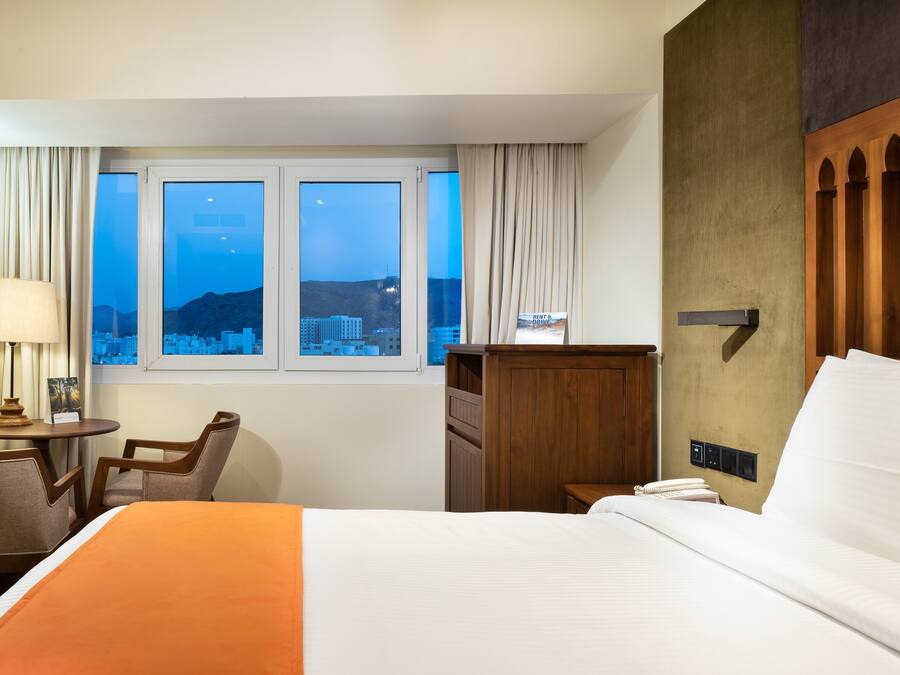 Habitación de hotel con cama, escritorio con lámpara, silla, ventana con cortinas y vista a un paisaje urbano al anochecer.