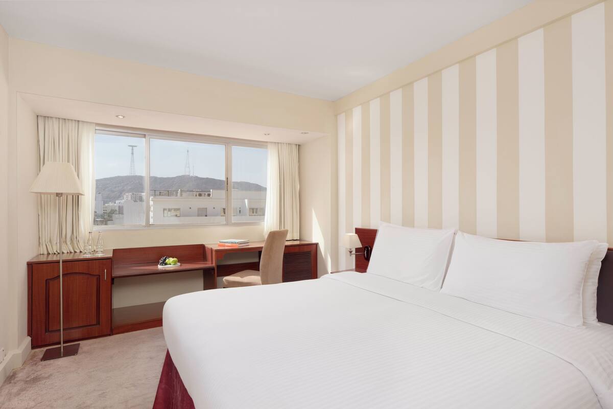A hotel room with a large bed, a desk, a chair, and a window with a city view.