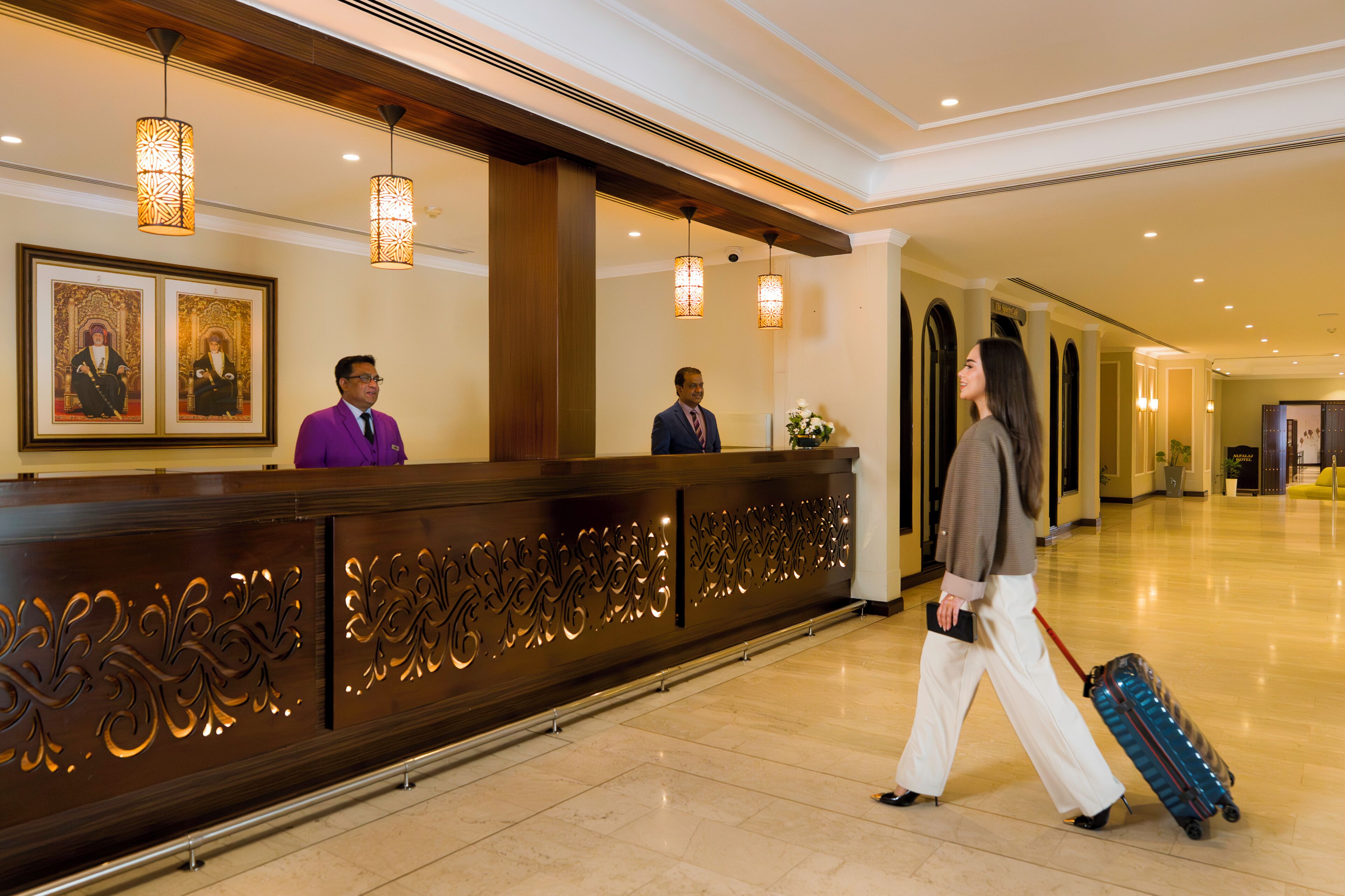 Zona de recepción de hotel con un mostrador de madera, dos recepcionistas y un huésped con maleta.