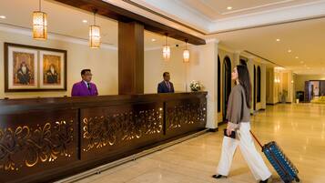 Zona de recepción de hotel con un mostrador de madera, dos recepcionistas y un huésped con maleta.
