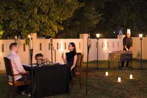 Un área de comedor al aire libre con una mesa dispuesta para dos personas, rodeada de velas y un chef en la estación de servicio.