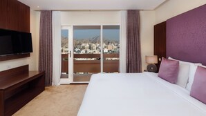 A hotel room with a bed, a TV, a balcony with a city view, and a window with curtains. - Al Falaj Hotel (Muscat)