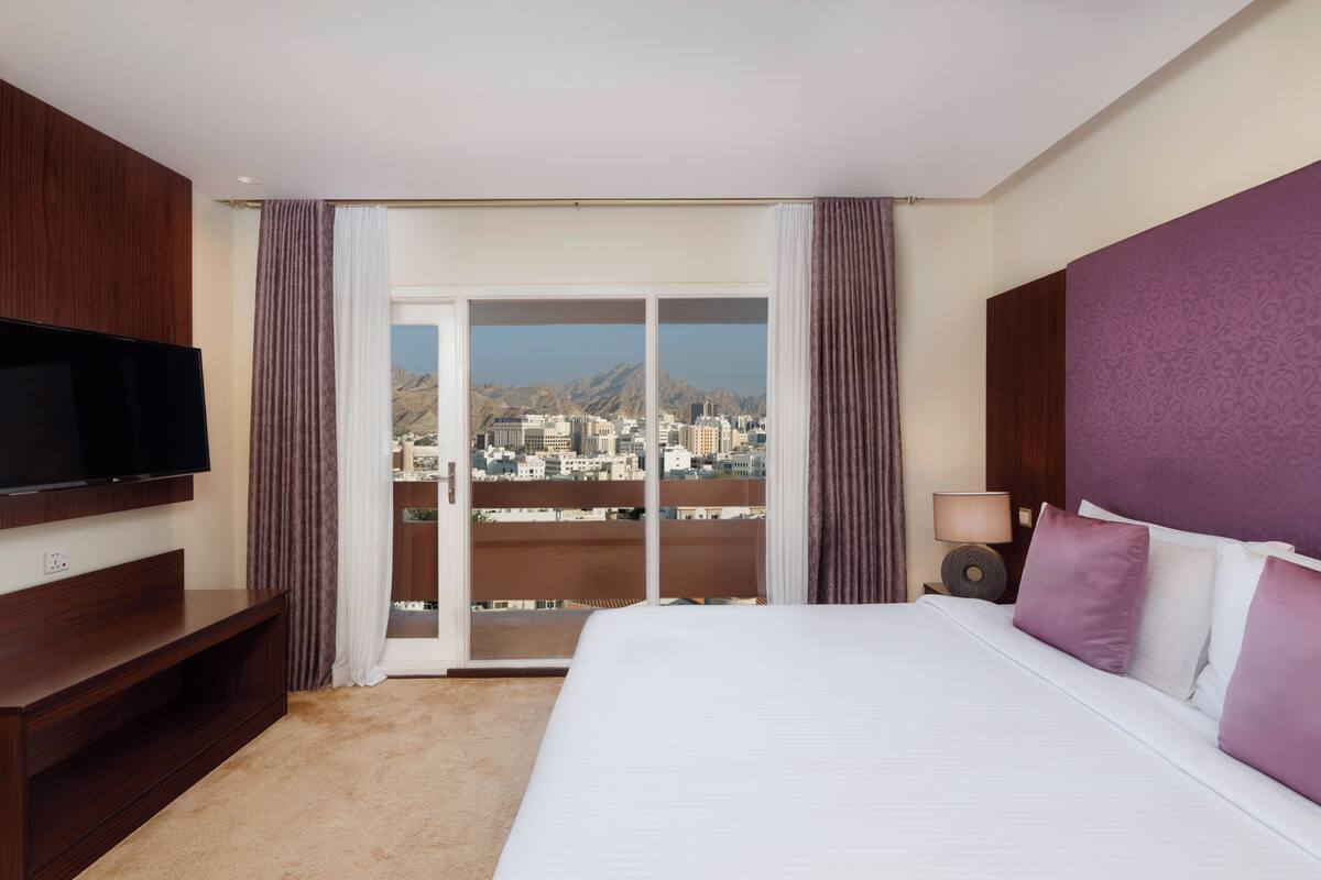 A hotel room with a bed, a TV, a balcony with a city view, and a window with curtains.