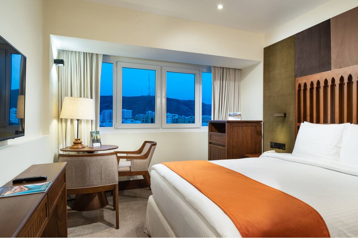 A hotel room with a large bed, a desk with a chair, a window with curtains, and a view of the cityscape.