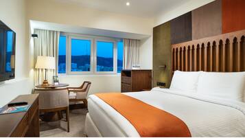 Habitación de hotel con una cama grande, un escritorio con silla, una ventana con cortinas y vistas a la ciudad.