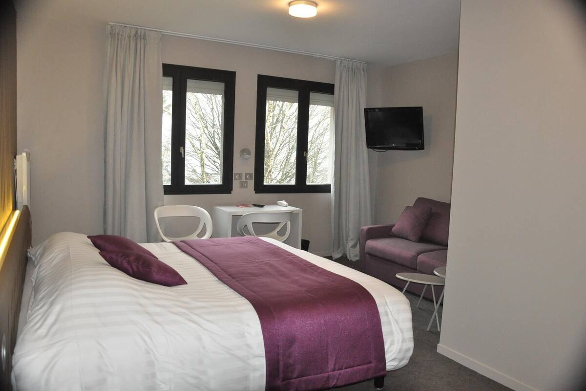 Een hotelkamer met een bed, een bureau, twee stoelen, een paarse bank en een aan de muur bevestigde televisie.