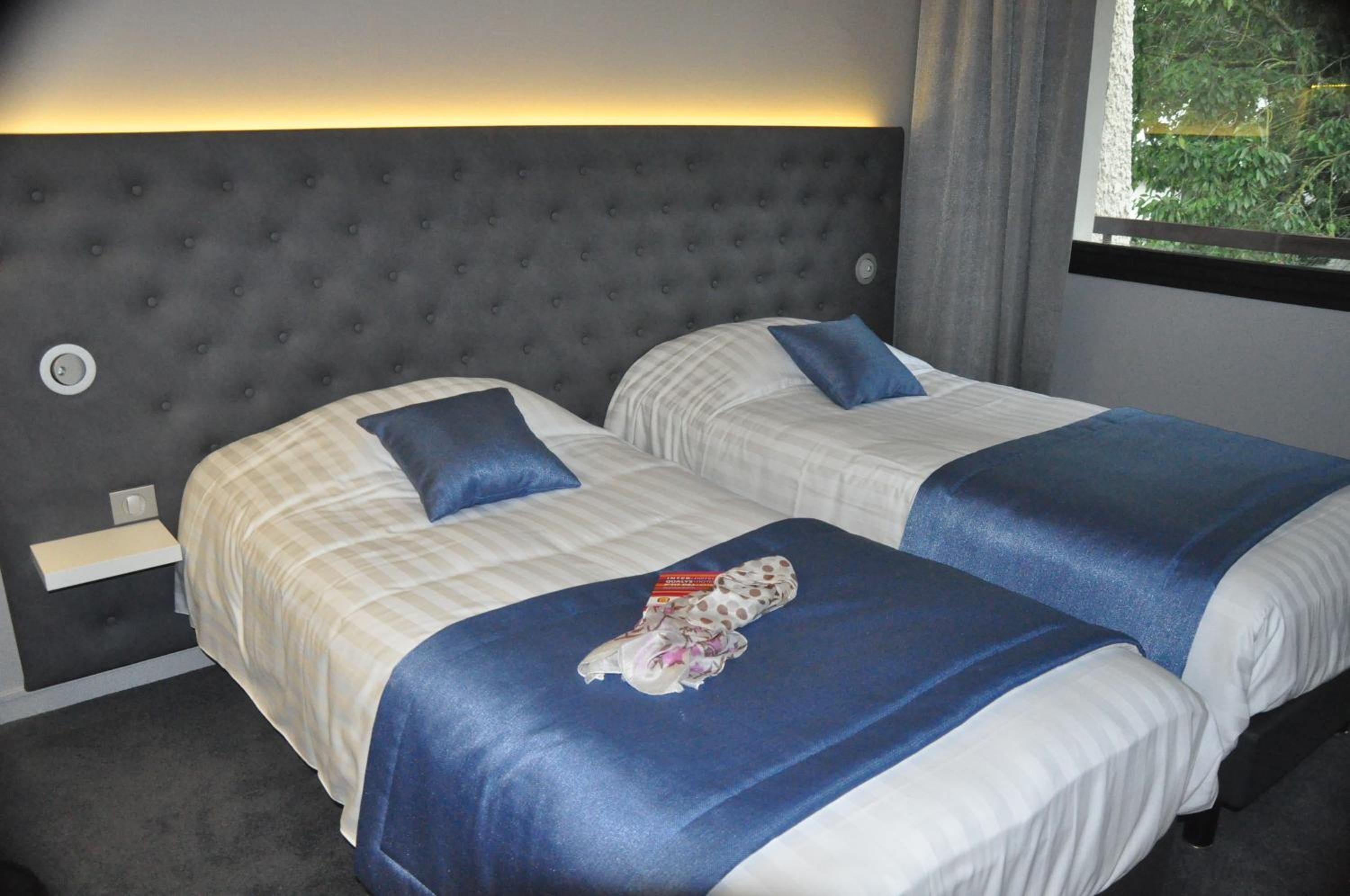 A hotel room with two beds, each with a blue bedspread and pillow, a grey tufted headboard, and a small bedside table.