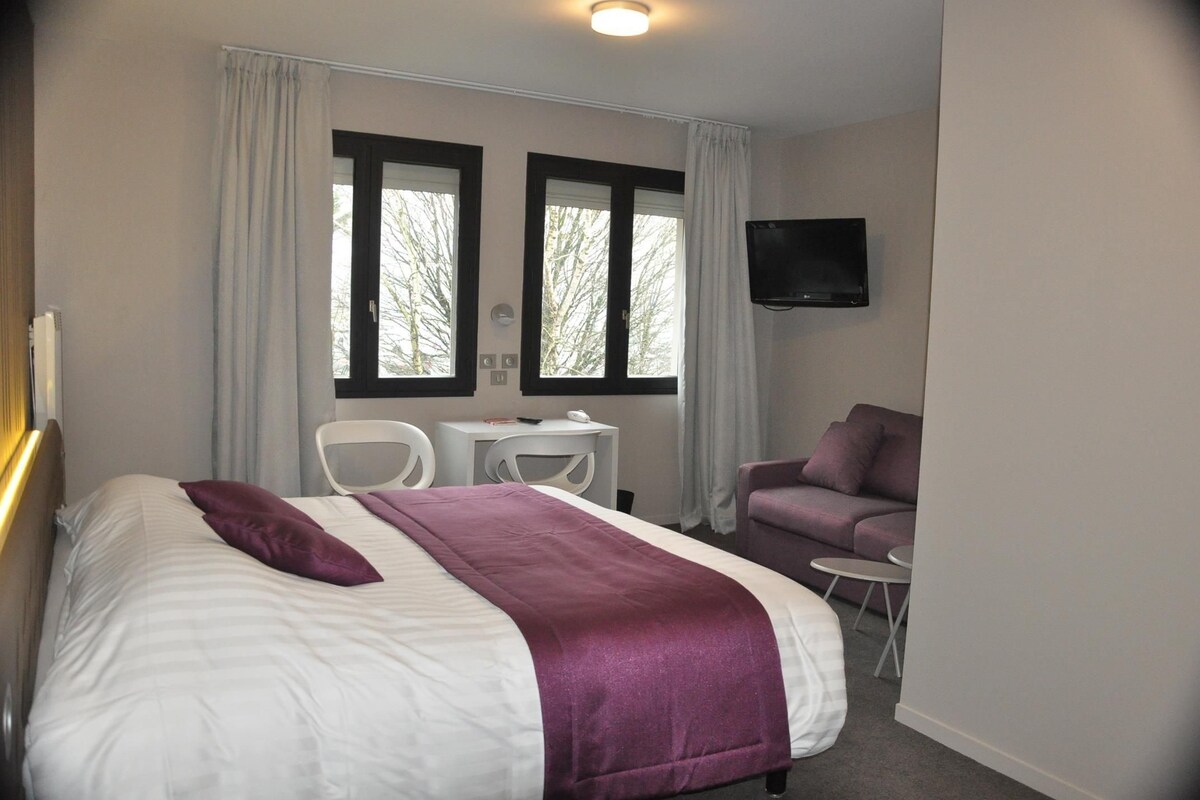 Une chambre d’hôtel avec un lit, un bureau, deux chaises, un canapé violet et un téléviseur fixé au mur.