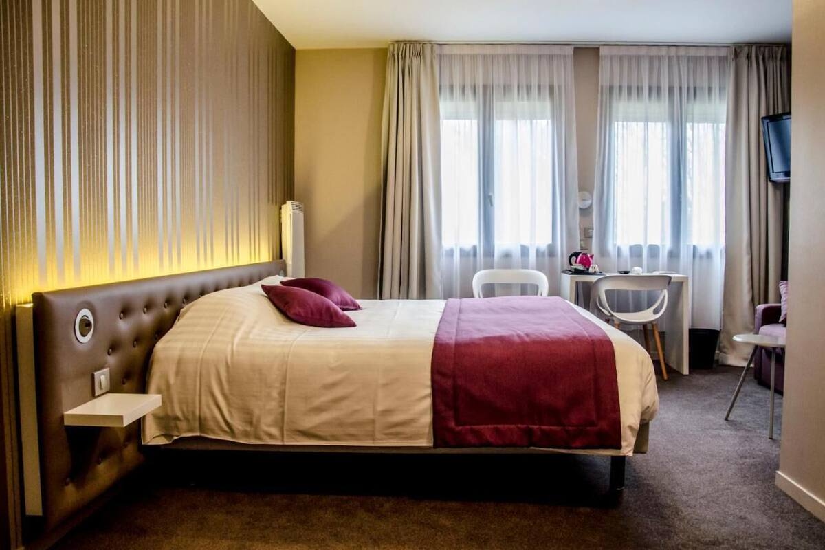 Hotelkamer met een groot bed, een bureau en een stoel. Een televisie hangt aan de muur en er is een raam met gordijnen.