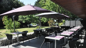 Un espace extérieur aménagé pour les repas, avec des tables et des chaises placées sous des parasols, le tout entouré de verdure.