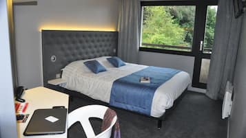 A hotel room with a large bed, a desk with a laptop, and a window with a view of greenery.