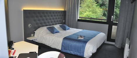 Een hotelkamer met een groot bed, een bureau met een laptop en een raam met uitzicht op groen.