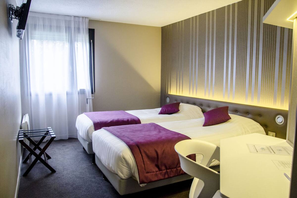Een hotelkamer met twee bedden, een kleine tafel, een stoel en een raam met dunne gordijnen.
