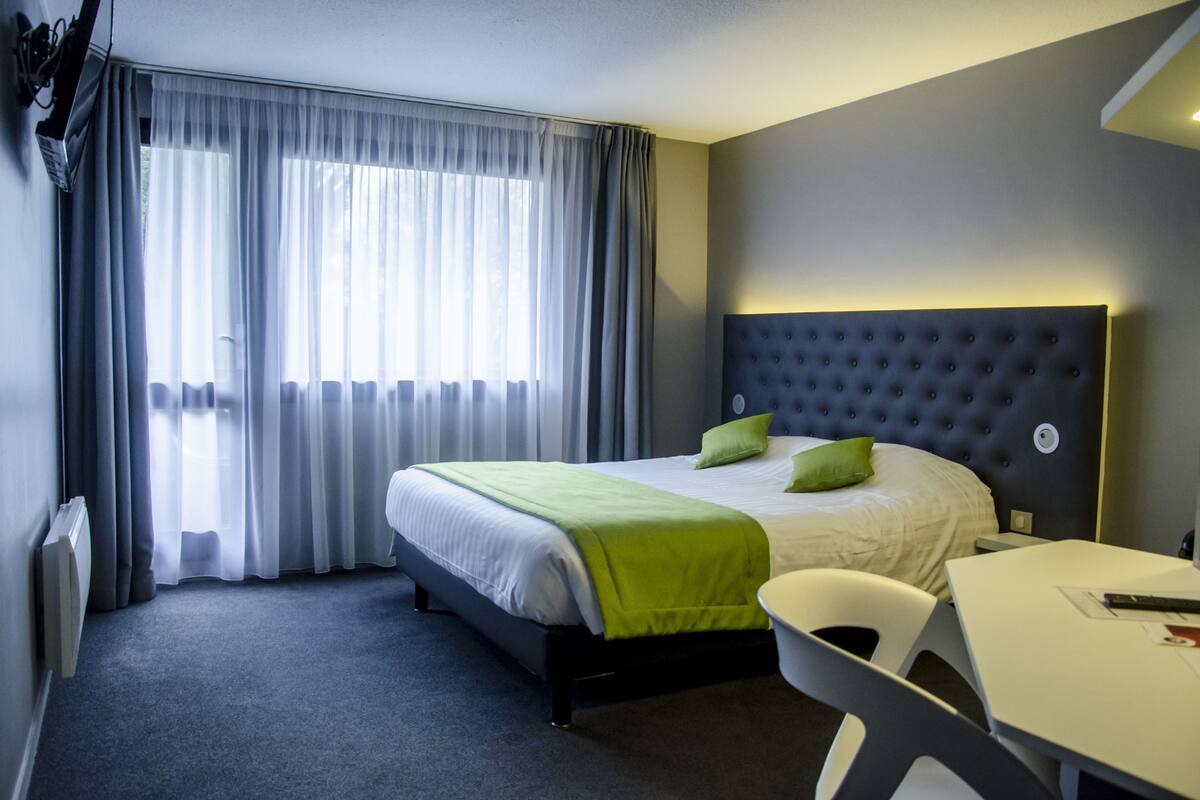 Een hotelkamer met een bed, een bureau, een stoel, een raam met gordijnen en een wandlamp.