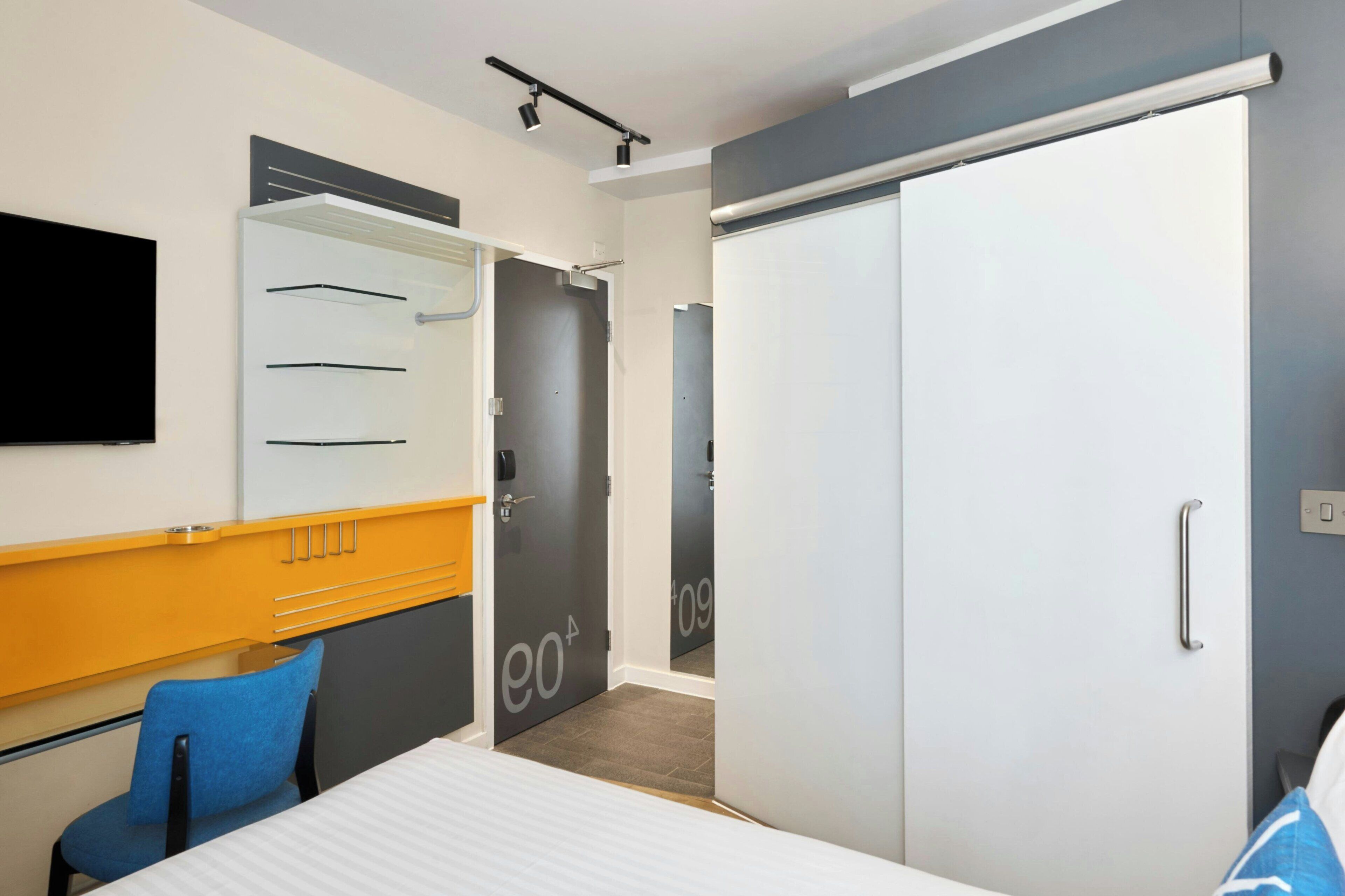 A modern hotel room with a bed, a desk, a chair, a wardrobe, and a television.