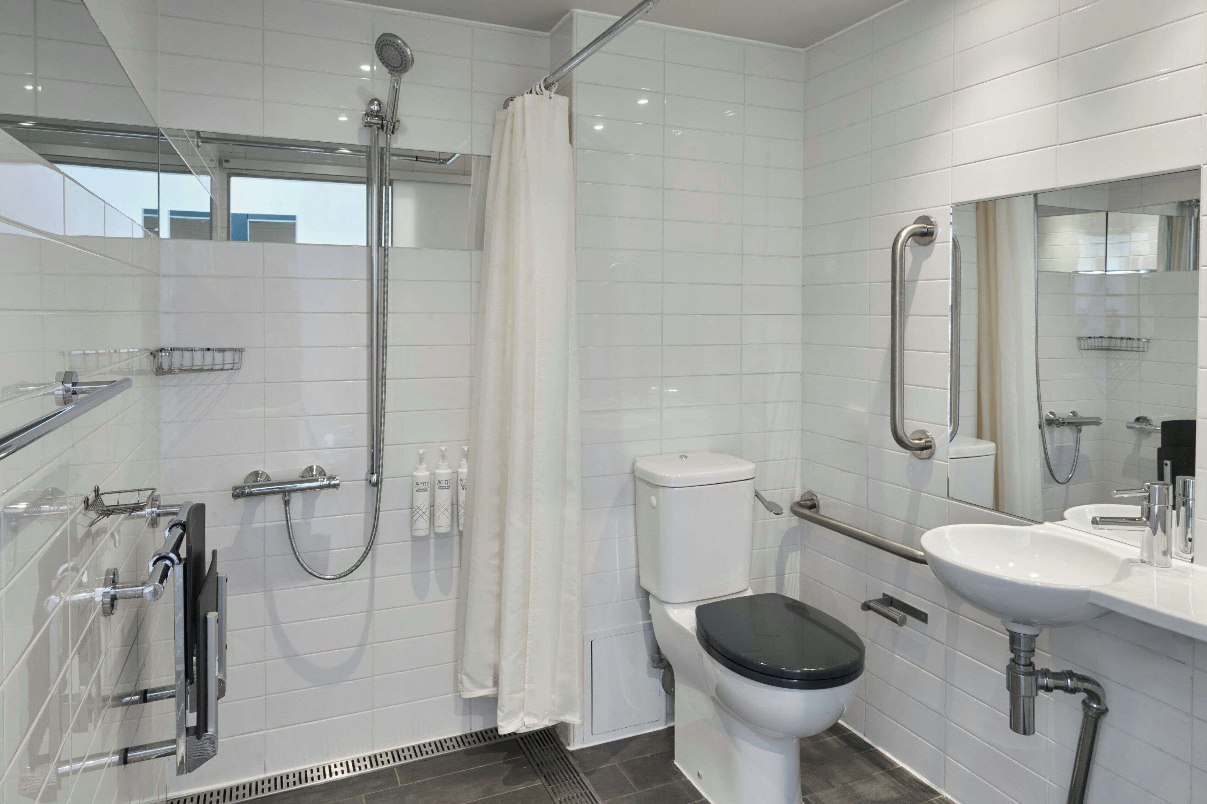 A modern bathroom with a shower, toilet, and sink, equipped with grab bars and a hand dryer.