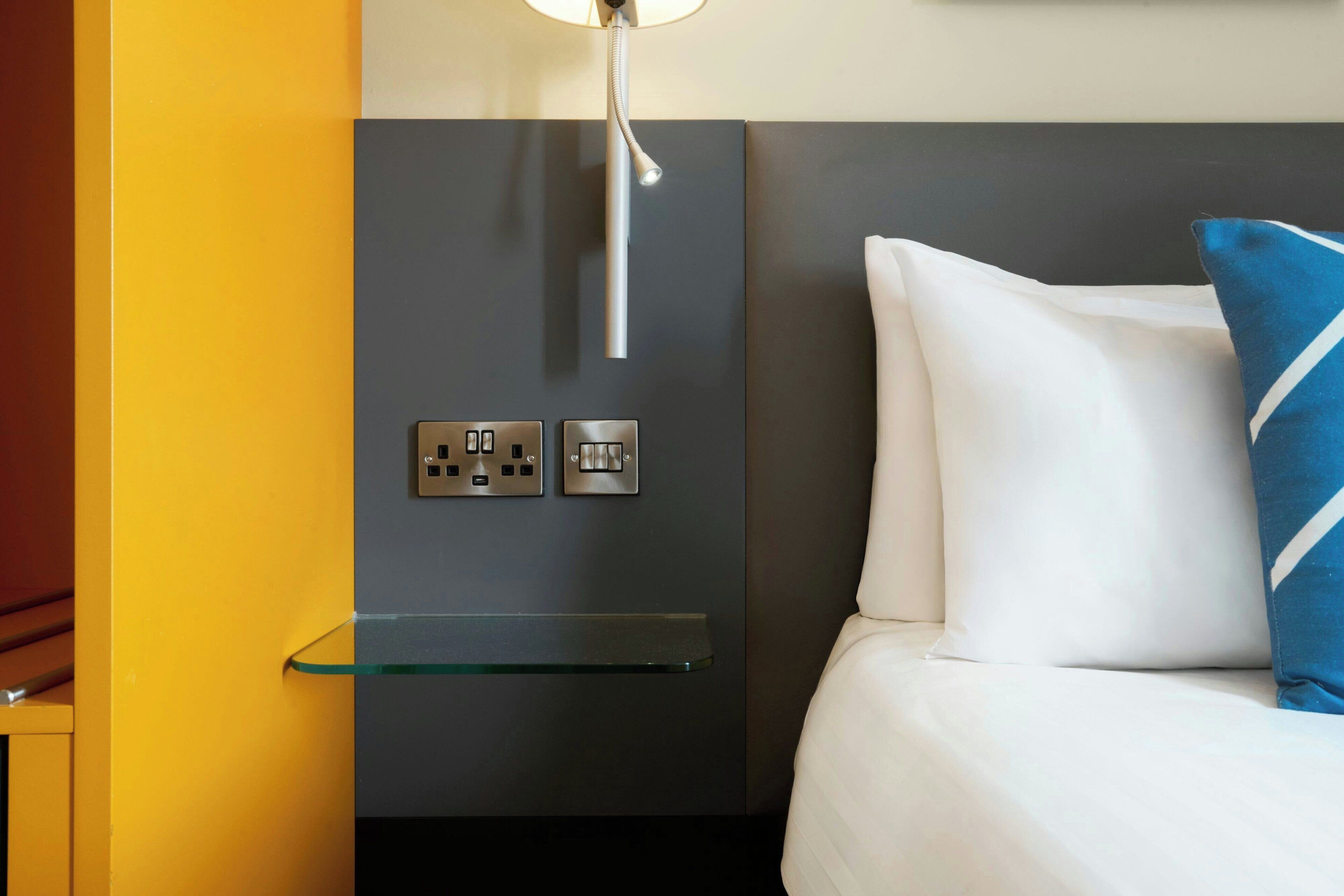 A modern hotel room with a glass shelf, a bed with white and blue pillows, and a wall-mounted light fixture.