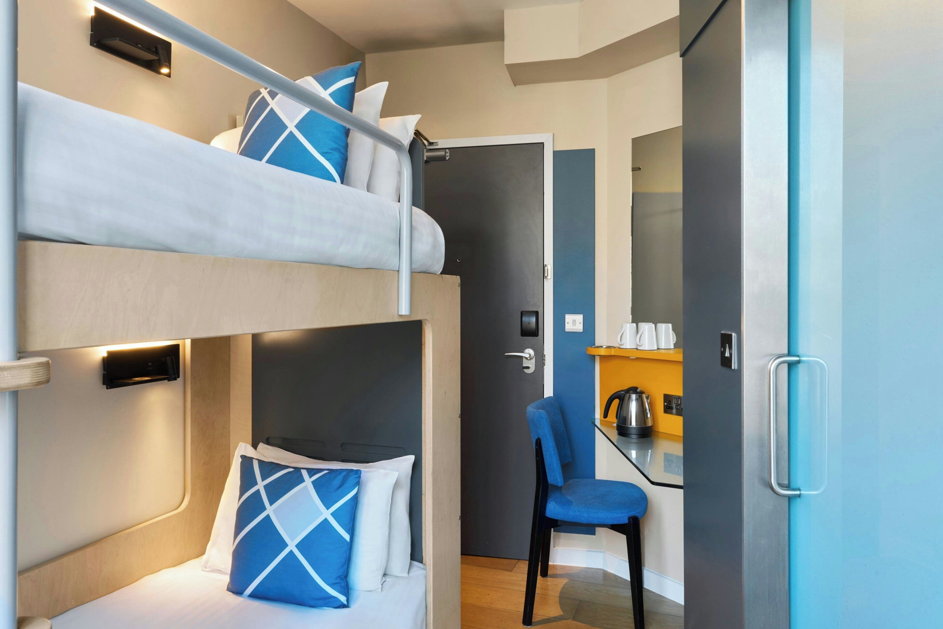 A bunk bed with pillows, a chair, a kitchen area with a kettle, and a door.