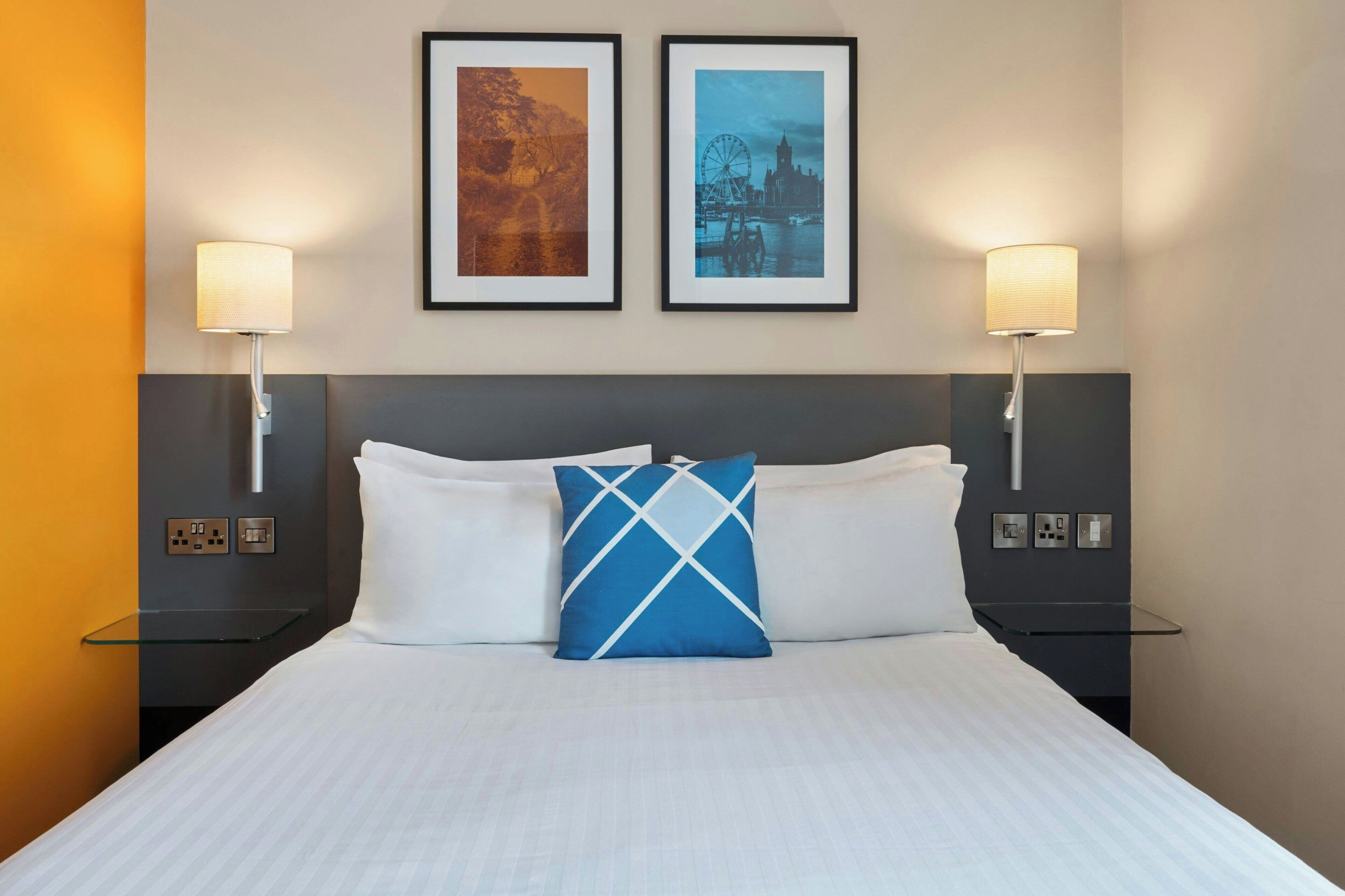 A hotel room with a large bed, two wall-mounted lamps, and two framed pictures above the bed.