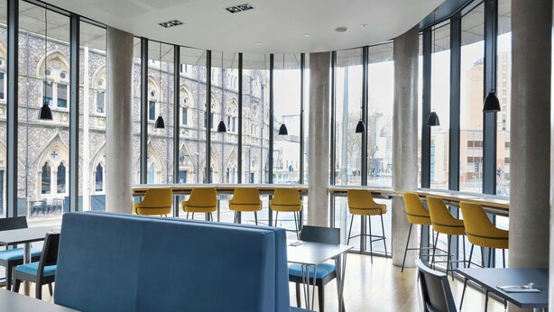 Un comedor moderno con asientos azules y sillas amarillas, amplias ventanas que ofrecen vistas de arquitectura histórica.