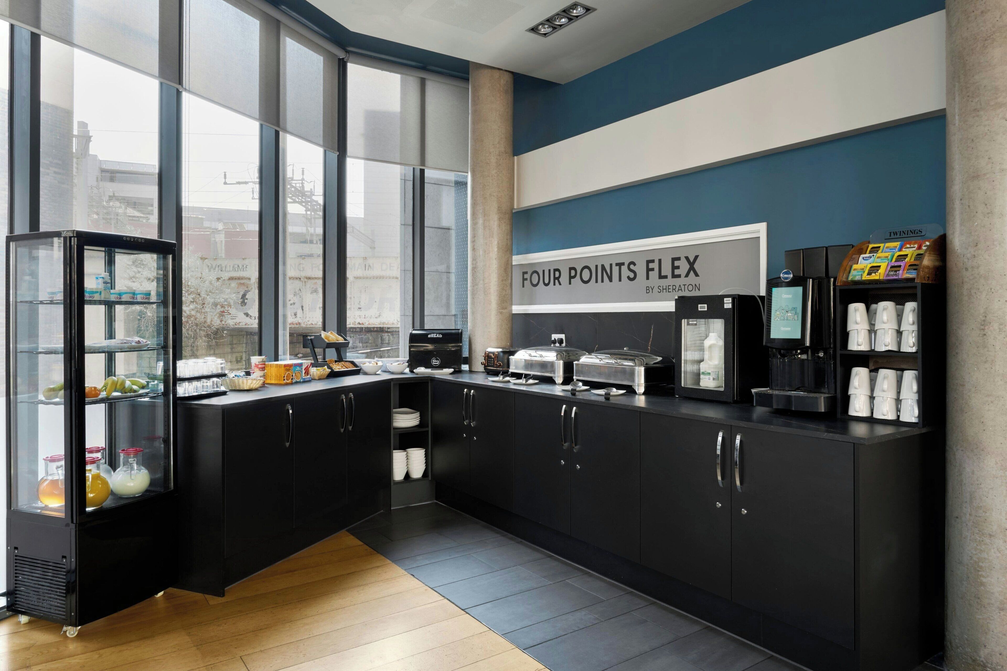 A modern kitchen with a sign reading 'FOUR POINTS FLEX BY SHERATON,' featuring a coffee machine, a bread maker, and a refrigerator.