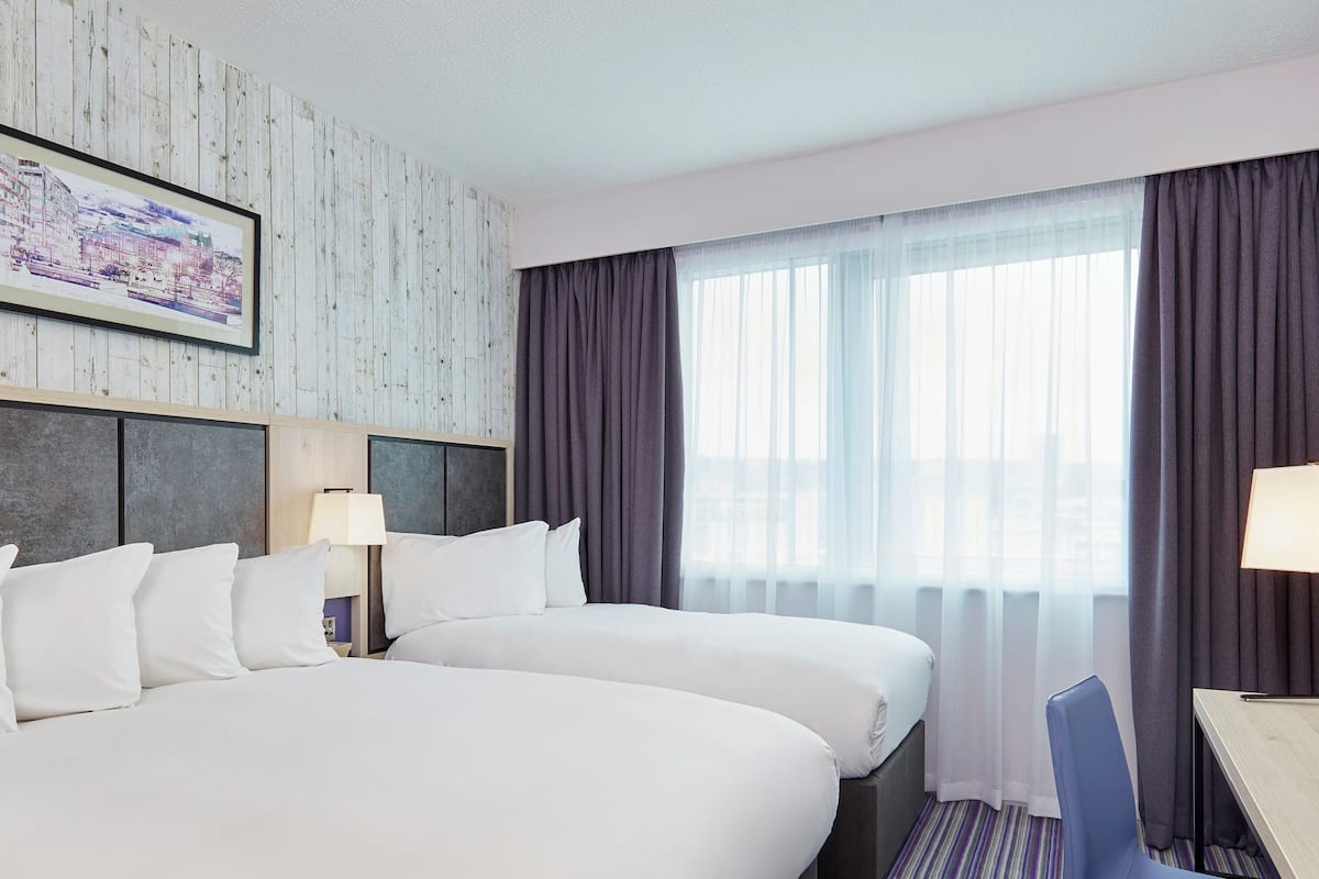 Habitación de hotel con dos camas, un ventanal con cortinas translúcidas, un cuadro con vista urbana en la pared y una pequeña mesa con una silla.