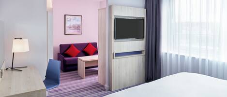 Une chambre d’hôtel avec un lit, un bureau avec une lampe, un canapé avec des coussins rouges, un téléviseur posé sur un meuble en bois et une fenêtre avec des rideaux légers.