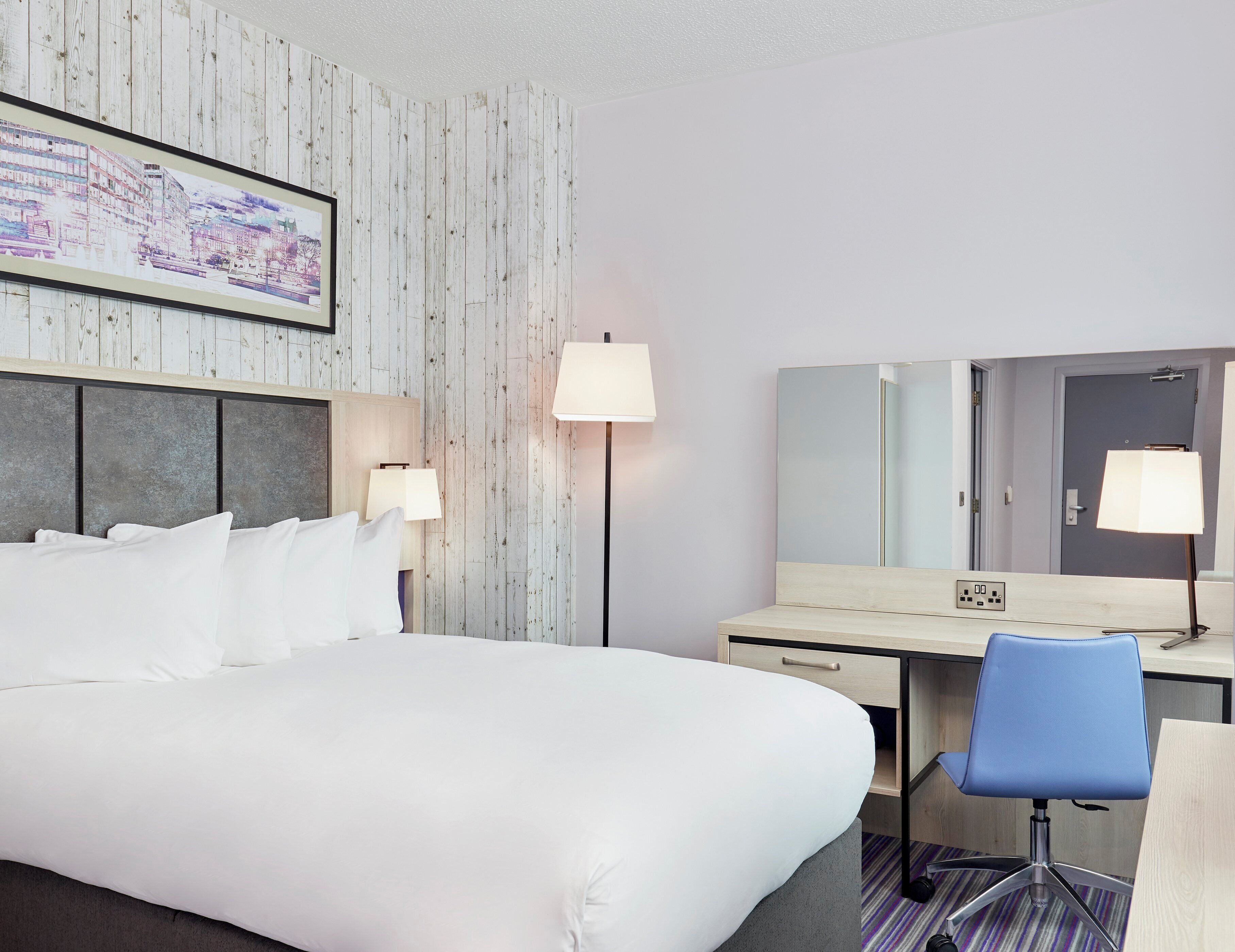 A hotel room with a bed, bedside lamps, a desk with a chair, a large mirror, and a wall-mounted TV.