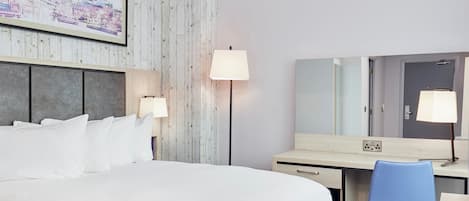 A hotel room with a bed, bedside lamps, a desk with a chair, a large mirror, and a wall-mounted TV.