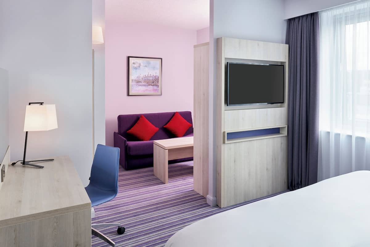 Habitación de hotel con cama, escritorio con lámpara, sofá con cojines rojos, televisor sobre un mueble de madera y ventana con cortinas traslúcidas.