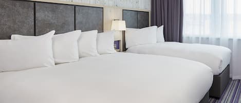 A hotel room with two beds, a nightstand with a lamp, and a view of the city through sheer curtains.