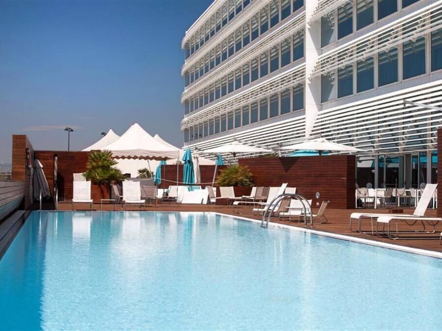 Una piscina con tumbonas y sombrillas, una terraza de madera y un edificio moderno con múltiples ventanas al fondo.