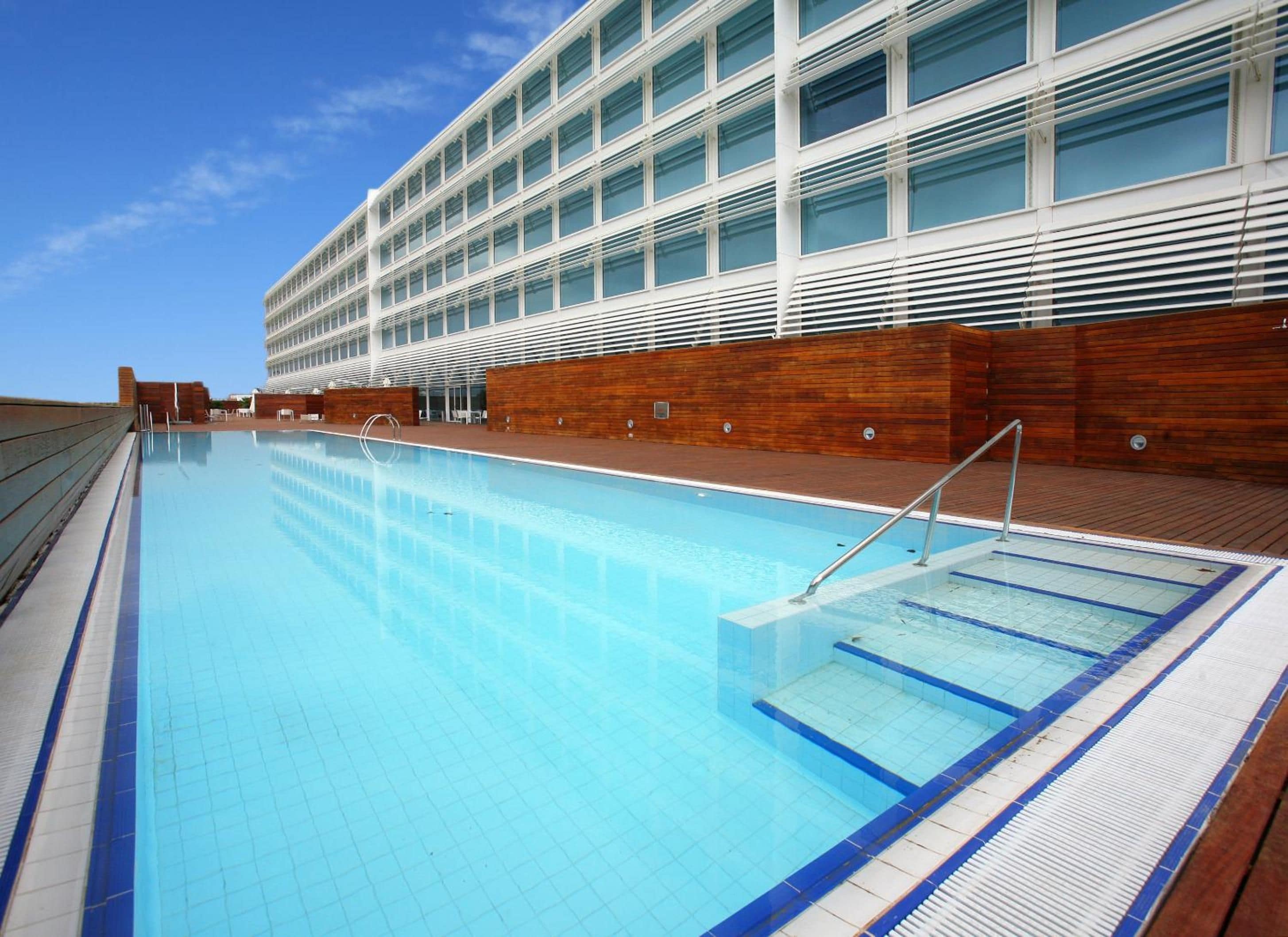 Una piscina con agua cristalina azul, rodeada por una terraza de madera y un edificio moderno con grandes ventanales.