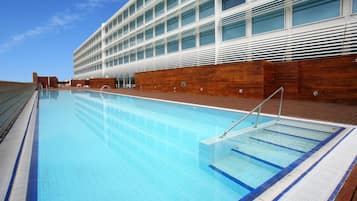 Una piscina con agua cristalina azul, rodeada por una terraza de madera y un edificio moderno con grandes ventanales.
