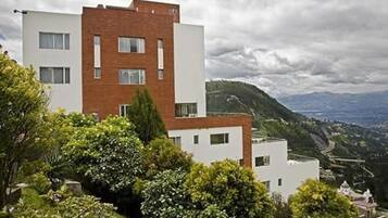 A modern two-story building with a flat roof and a brick facade, surrounded by lush greenery and overlooking a hilly landscape.