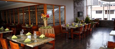 A restaurant with wooden tables and chairs, a large vase with flowers, and a view of buildings through large windows.