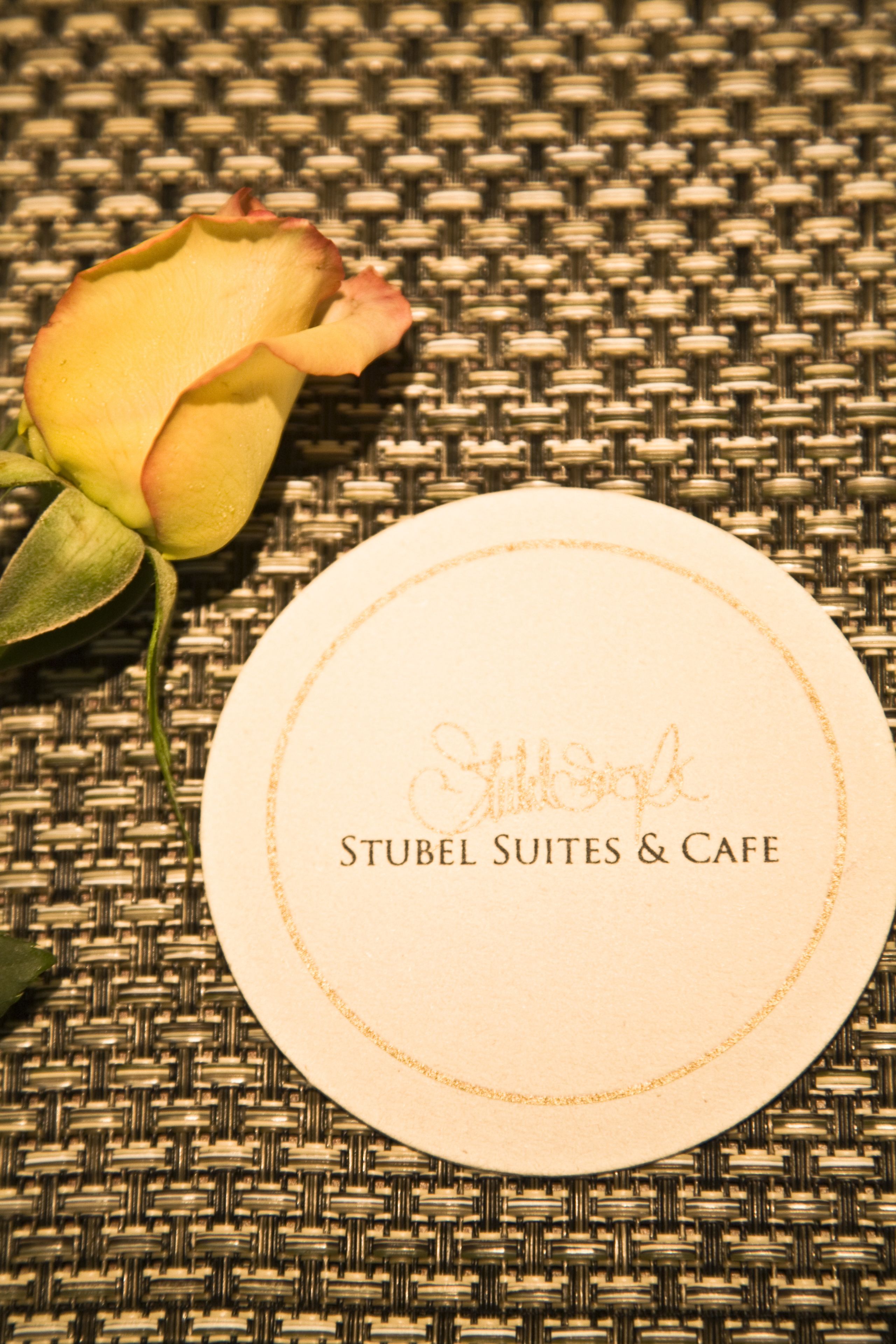 A round white card with "Stubel Suites & Cafe" embossed on it, placed on a woven surface with a single rose beside it.