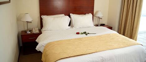 Una habitación de hotel con una cama grande, dos mesitas de noche con lámparas, un cabecero de madera y una rosa roja sobre la cama.