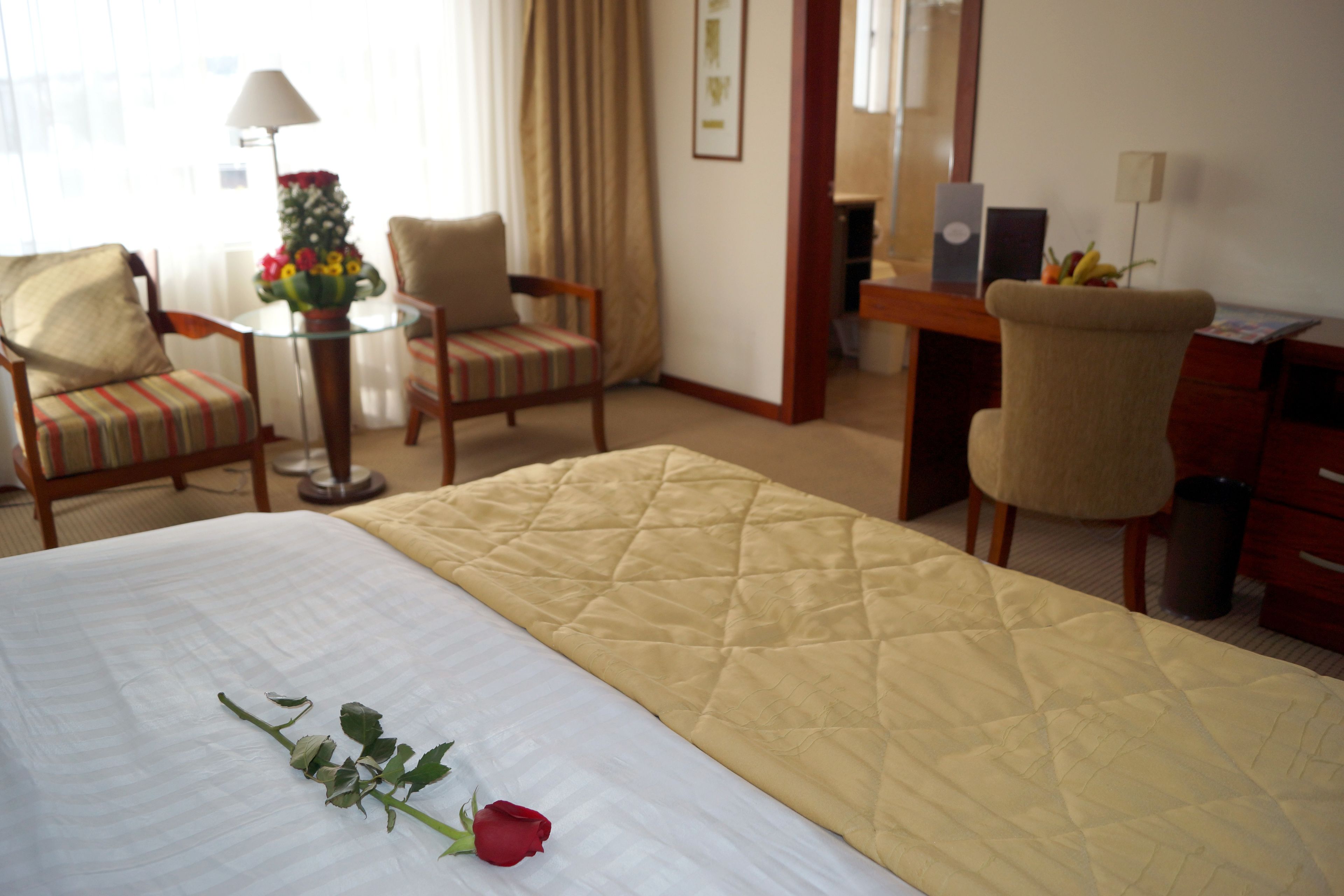 Una habitación de hotel con cama, dos sillones, un escritorio, una lámpara y una mesita con flores.