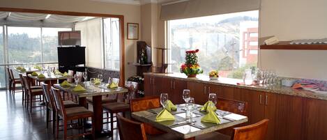 A dining area with multiple tables set for guests, wooden chairs, and a large window offering a scenic view.