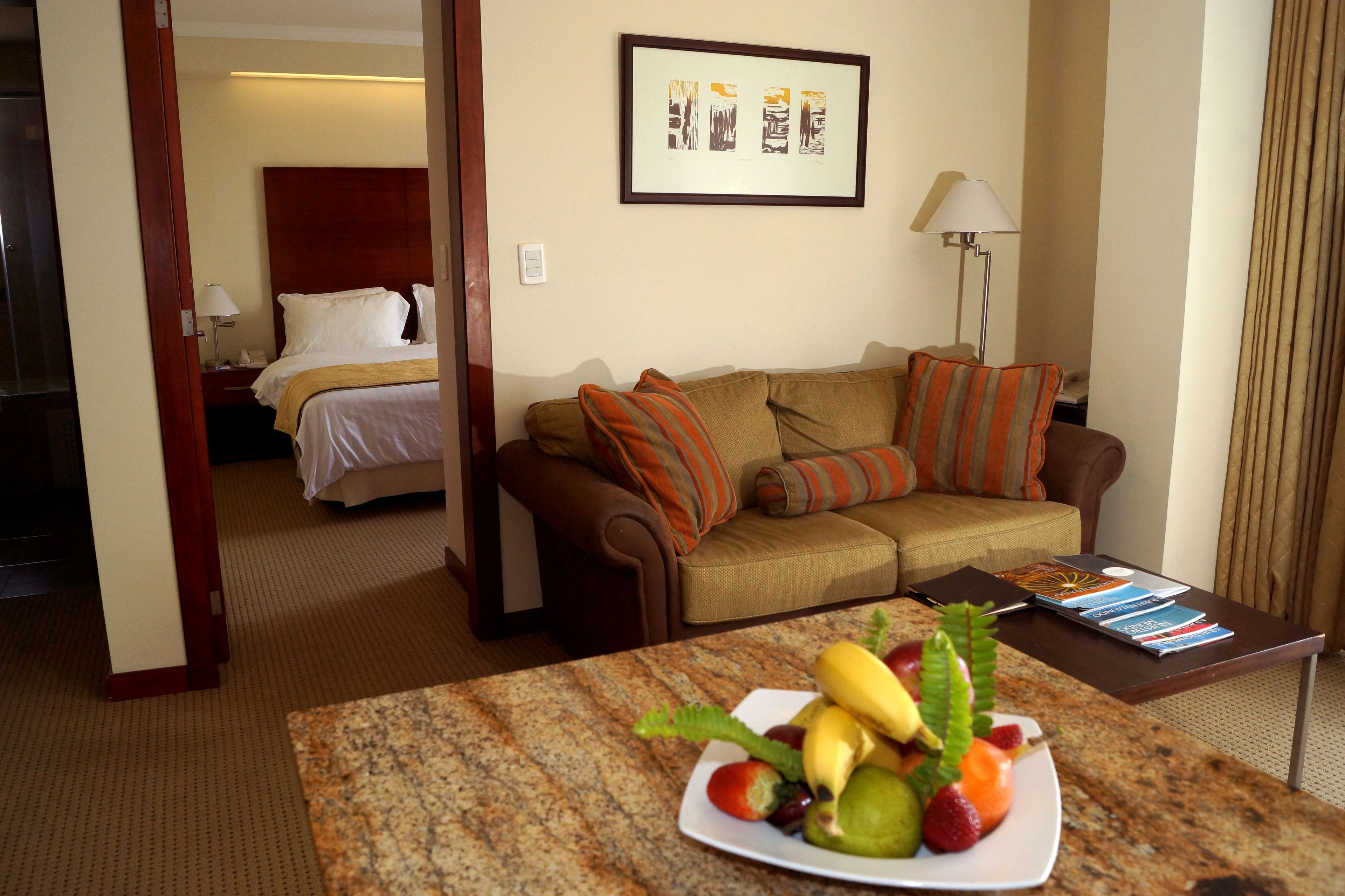 Une chambre d’hôtel avec un lit, un canapé, une table basse avec des fruits et un tableau encadré au mur.
