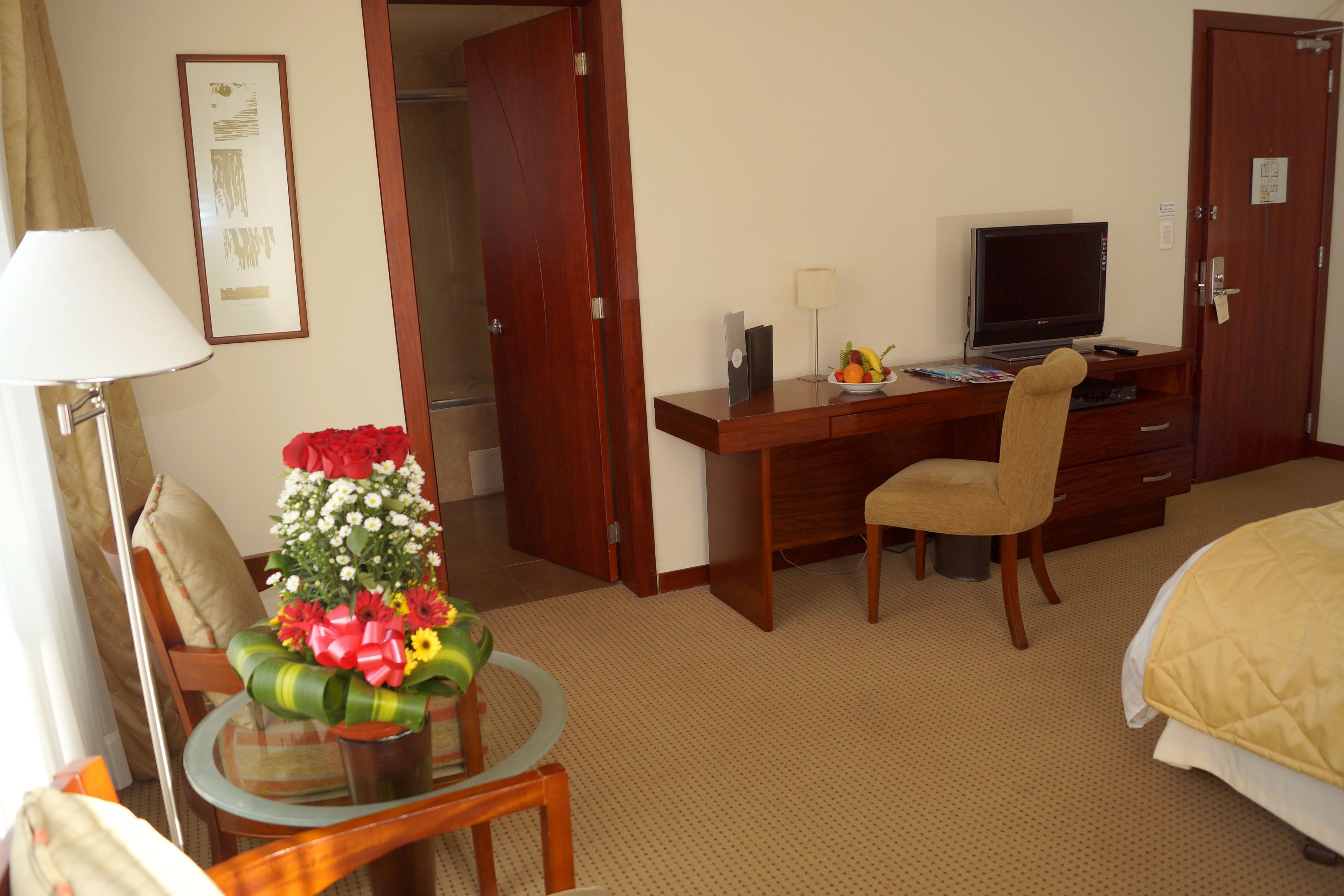 Una habitación de hotel con una cama, un escritorio con computadora, una silla, una mesita con flores y una lámpara.